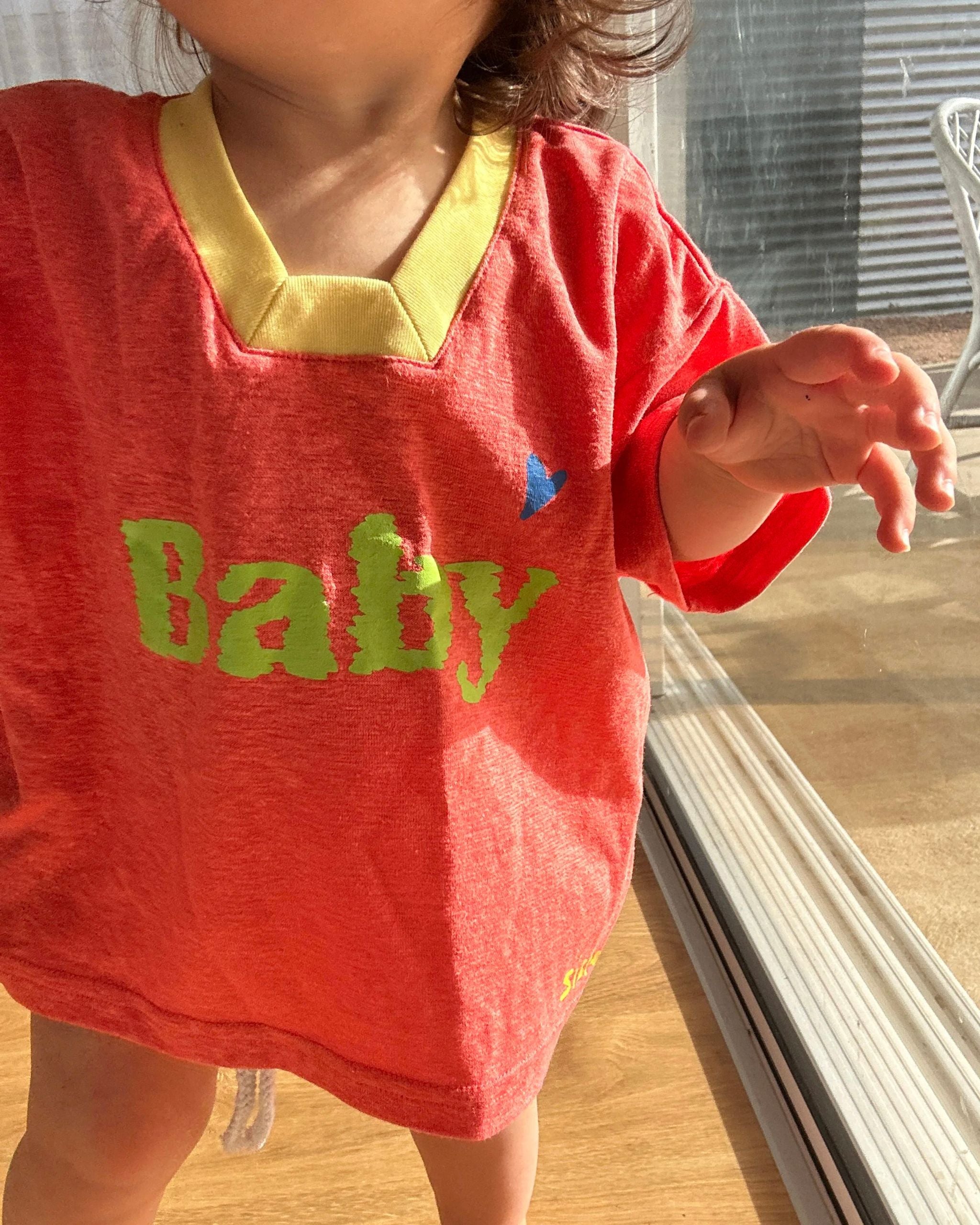 Child showing a colorful outfit while wearing an orange-peach hemp and tencel kids tee with ‘baby sister’ text by Yolky Club