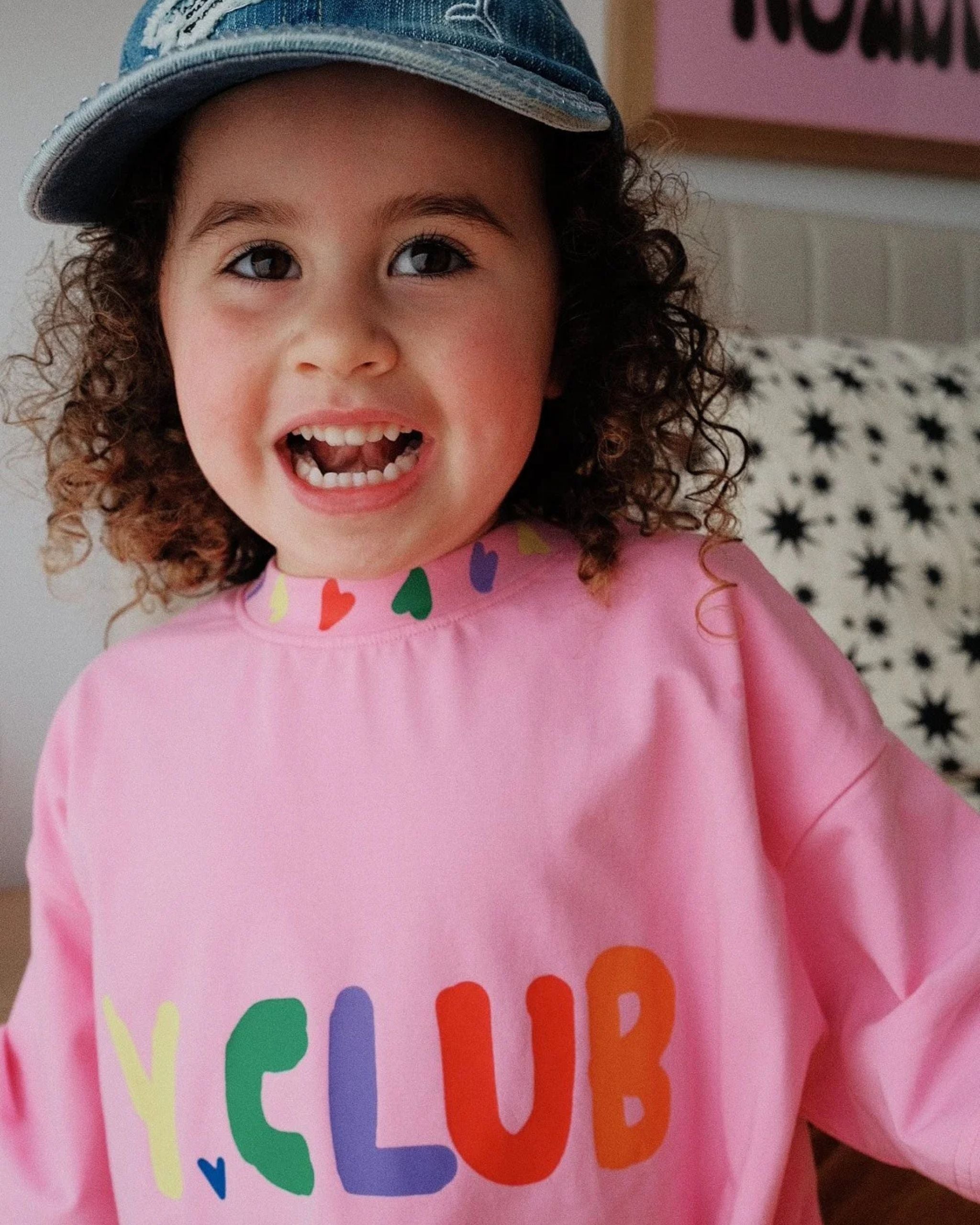 Young girl smiling while wearing a pink Y.CLUB kids tee with rainbow lettering and heart details by Yolky Club